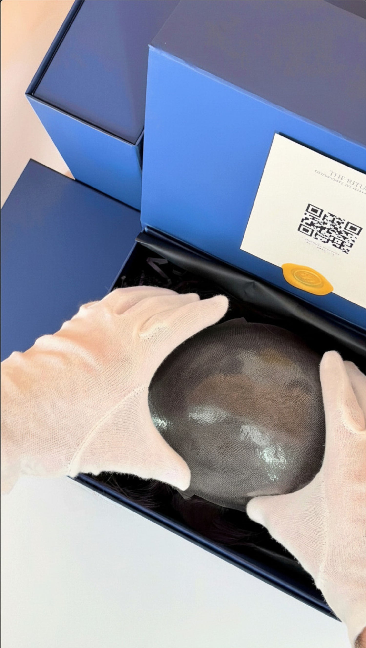 Person wearing gloves holding a thin skin hair system for men in front of a blue box with a QR code.
